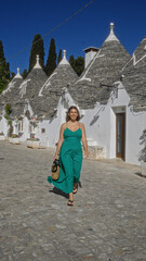 Wall Mural - A beautiful young hispanic woman walking down a charming street lined with traditional trulli houses in the old town of alberobello, puglia, italy, wearing a green dress and carrying a purse.