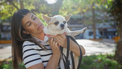 Wall Mural - A young hispanic woman holds a chihuahua dog in an urban park setting, conveying companionship and leisure outdoors.