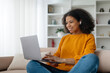 © Anastasiya - African American Female Freelancer Using Laptop And Wearing Wireless Earbuds Working Online On Computer Sitting on the sofa in the living room At Home. Freelance Lifestyle Concept. High quality photo