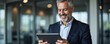 © schiers_images - A mature businessman smiling while interacting with a digital tablet in a contemporary office environment, highlighting professional success and modern technology use.