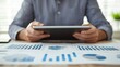 © pattaphorn - A person holding a tablet, analyzing various blue graphs and charts spread out on a table in a well-lit environment.