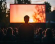 © Tanakrit - A silhouetted figure watches a vibrant sunset on a large outdoor screen, surrounded by an audience in a serene setting.