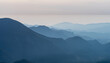 © Trinitro - Early morning mist envelops rolling hills and mountains in a serene landscape at dawn, showcasing layers of blue and gray hues stretching into the horizon