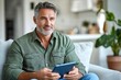 © tiger - Middle aged man relaxing on couch using tablet in modern living room with casual attire and thoughtful expression