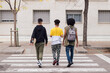 © Raul Mellado - rear view of three diverse teen friends with backpack and skateboard crossing road on crosswalk, concept of youth and urban lifestyle