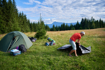 Wall Mural - Man hiker set up tourist tent on grassy hillside. Pine trees and distant mountains under partly cloudy sky.