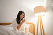© Odua Images - A young woman is happily using her smartphone in a cozy bedroom, creating a peaceful atmosphere