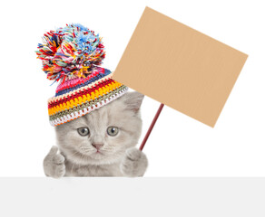 Aufkleber - Cute kitten wearing a warm hat with pompon looking above blank banner, showing thumbs up gesture and showing blank placard on wooden stick. isolated on white background