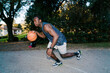 © Studio Marmellata - Black young man wearing athletic shorts and a tank top dribbles a basketball on an outdoor court surrounded by trees and nearby residential buildings during a sunny afternoon. flash light motion blur