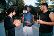 © Studio Marmellata - Three basketball players stand on an outdoor court, one holding a basketball, as they talk and smile, with trees and a basketball hoop in the background.