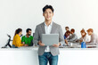 © Irina B - Asian man holding a laptop, standing in a bright collaborative workspace with colleagues working together in the background, emphasizing teamwork and innovation..
