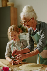 Elderly woman teaching young girl baking, both enjoying shared activity in cozy kitchen setting. Senior and child wearing matching aprons, surrounded by kitchen tools and ingredients