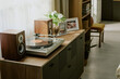 © AnnaStills - Turntable set on wooden cabinet beside photo frames and potted plants with sunlight streaming through a nearby window, creating a serene and nostalgic ambiance