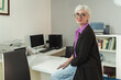 © Lomb - Stylish businesswoman with white pixie hair and glasses smiling in modern office workspace with computer