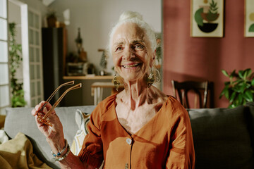 Wall Mural - Elderly woman standing in living room, smiling while holding glasses. Cozy interior features plants, artwork, and modern furniture creating homey atmosphere