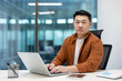 © Liubomir - Asian man businessman using laptop at office desk, looking confident. Surroundings include smartphone and notes. Modern workspace evokes professionalism, focus, determination, productivity.
