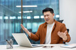 © Liubomir - Asian man businessman showing frustration while working on laptop in office. Expressing stress and confusion with hands up and bewildered expression, phone on desk.
