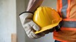 © Nata - A construction worker wearing a yellow hard hat and orange safety vest. The worker is holding a pair of gloves in his right hand