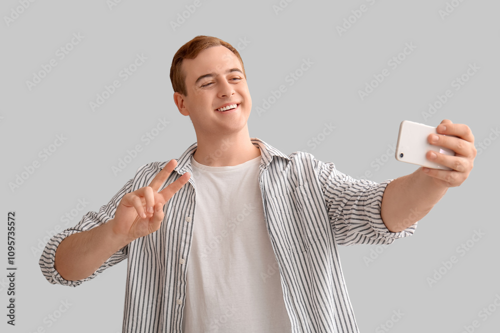 Young man with mobile phone showing victory gesture and taking selfie on light background