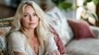 © Giovanni - Middle-aged blonde woman with long hair wearing a light-colored outfit, relaxing on a patterned sofa surrounded by plants and cushions.