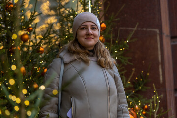 Portrait of serene adult caucasian woman in grey jacket and hat standing by green decorated Christmas tree on city street in a cold cloudy day. Soft focus. People and winter holidays theme.