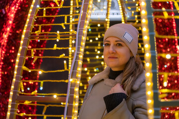 Portrait of serene adult caucasian woman in grey jacket and hat standing by illuminated orange and red Christmas street decoration on night winter city street. Soft focus. People and holidays theme.