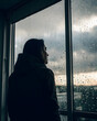 © JidapaDesign - figure facing away, looking out rain soaked window, reflecting on life. atmosphere is contemplative and serene, with raindrops adding sense of melancholy