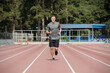 © pressmaster - Athletic man jogging on outdoor running track in well-maintained park surrounded by tall evergreen trees, wearing casual sports attire