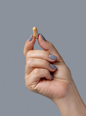 female hand holding a yellow capsule with fingers closeup against grey, healthcare concept