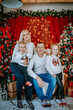 © Raivo - A cheerful family of four poses in a cozy holiday setting with Christmas trees, festive lights, and gift boxes, creating a warm seasonal atmosphere..