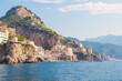 The photo captures a sunny view of Amalfi, showcasing the town's colorful buildings, steep cliffs, and vibrant coastline under a clear blue sky on a bright day.