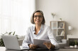 © fizkes - Happy successful business manager woman in glasses doing paperwork accounting job at home, working on freelance project at laptop, holding paper financial reports. Home portrait