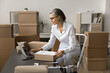 © fizkes - Mature grey haired businesswoman preparing box for shipping, transportation, sending to customer, filling up parcel card, typing on laptop, sitting on stepladder at workplace in storage room
