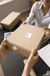 © fizkes - Internet shop storage worker giving cardboard box to female customer over workplace table. Client woman getting parcel in shipping office, taking package with bar code. Cropped shot of hands