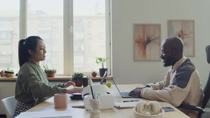 Medium side slowmo shot of happy Chinese female and African American male software developers sitting opposite in office, writing code on laptops, giving high fives on successful program test