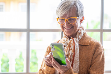 Wall Mural - Portrait of happy gray haired modern senior woman in yellow using smartphone close the window.