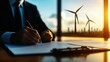© nattapon98 - green energy alternative policy Concept. A professional man signs a document, with wind turbines visible outside a modern office window, symbolizing renewable energy and sustainability.