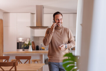 Wall Mural - Happy mid adult man looking through window and discussing over smart phone while standing at home