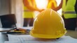 © Prapan - a bright yellow safety helmet placed on a modern office desk, with blueprints and project plans scattered around. In the background,