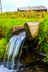 Natural water spring flowing from wooden pipe in green countryside with rustic farm buildings in background showing rural water system