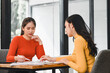 © kenchiro168 - Two women engaged in discussion while reviewing documents at table. One woman wears orange sweater, and other wears yellow sweater, showcasing collaborative atmosphere