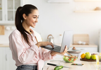 Aufkleber - Searching For New Recipes. Cheerful smiling young woman holding and using laptop, typing on keyboard, cooking fresh salad at home in modern kitchen, looking at screen, free copy space