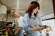© AnnaStills - Low angle shot of Asian female cook checking new orders in app on tablet while Caucasian male chef preparing hot dogs in blurred background