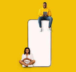 Poster - Great App. Excited Black Couple Sitting On Big Smartphone With Blank White Screen Using Gadgets, Cheerful Guy And Lady Chatting On Social Media, Yellow Orange Wall, Mock Up. Modern Communication