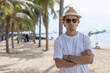 © eakgrungenerd - Handsome attractive man with sunglasses brimmed hat white T-shirt arm cross on tropical beach standing under palm tree in vacation summer travel.