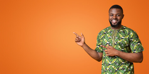 Wall Mural - Positive black man in authentic african clothes pointing at copy space over yellow studio background, panorama