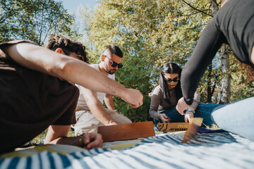 Canvas Print - Friends enjoy a strategic board game during a sunny picnic in a park, surrounded by lush greenery. The group is engaged and relaxed, highlighting teamwork and leisure.