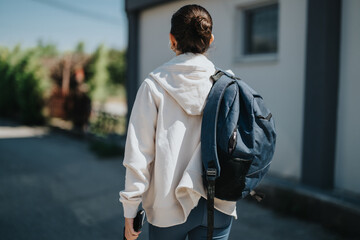 A young girl is walking outside, wearing a white hoodie and carrying a blue backpack. The setting appears urban and casual, conveying a sense of independence and youth.