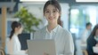 © Ganesha88 - Smiling Japanese businesswoman standing in an office, holding her laptop. She is wearing casual clothes, and other people working behind her are blurred.,generated ai