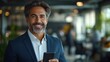 © jajang - A smiling man in a suit holds a smartphone in a modern office environment.